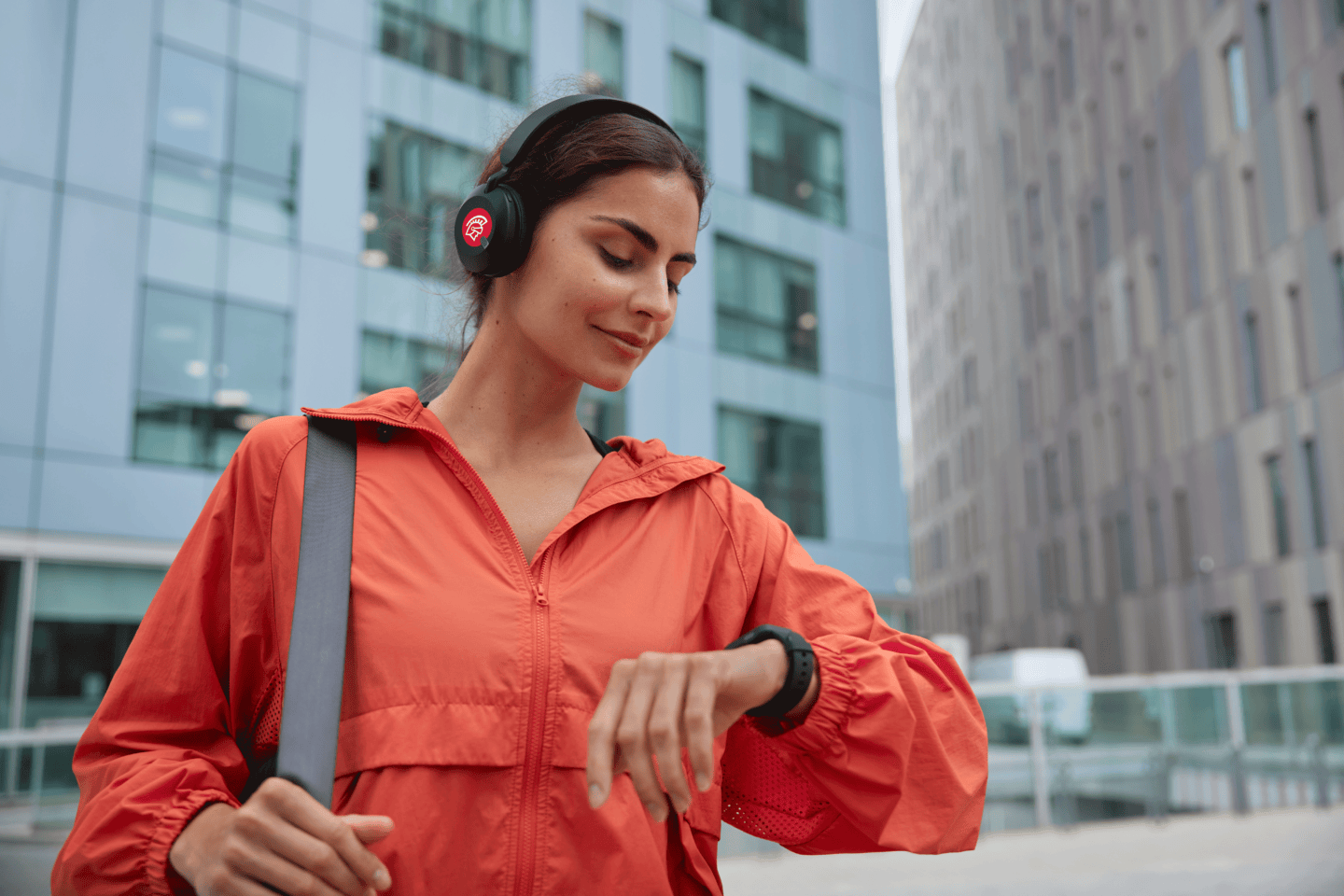 A woman listens to music with Safeguard branded headphones while looking down at her watch and standing in the middle of a city.