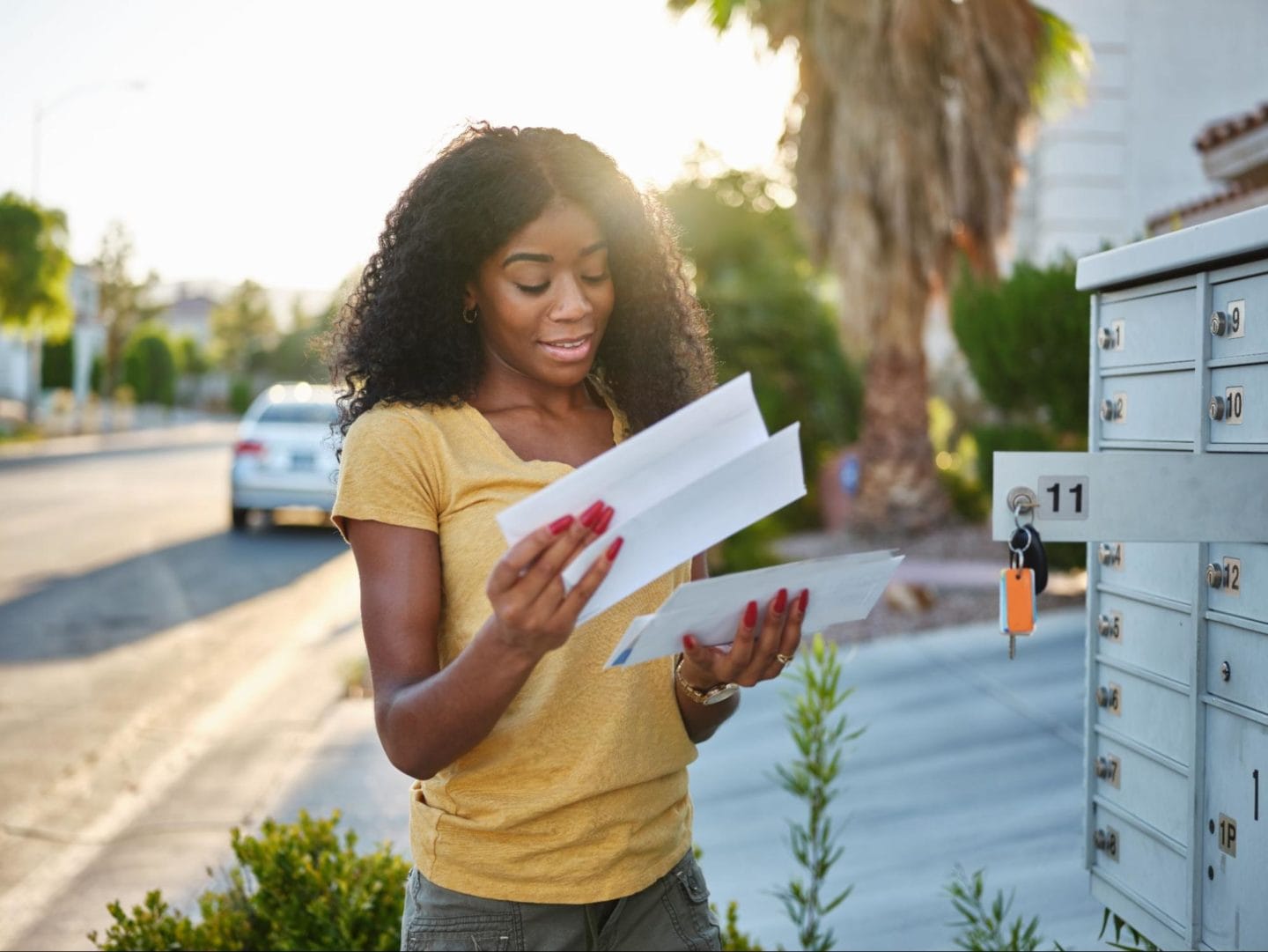 A picture of a young woman holding white envelopes and reading her mail after receiving it from her mailbox.