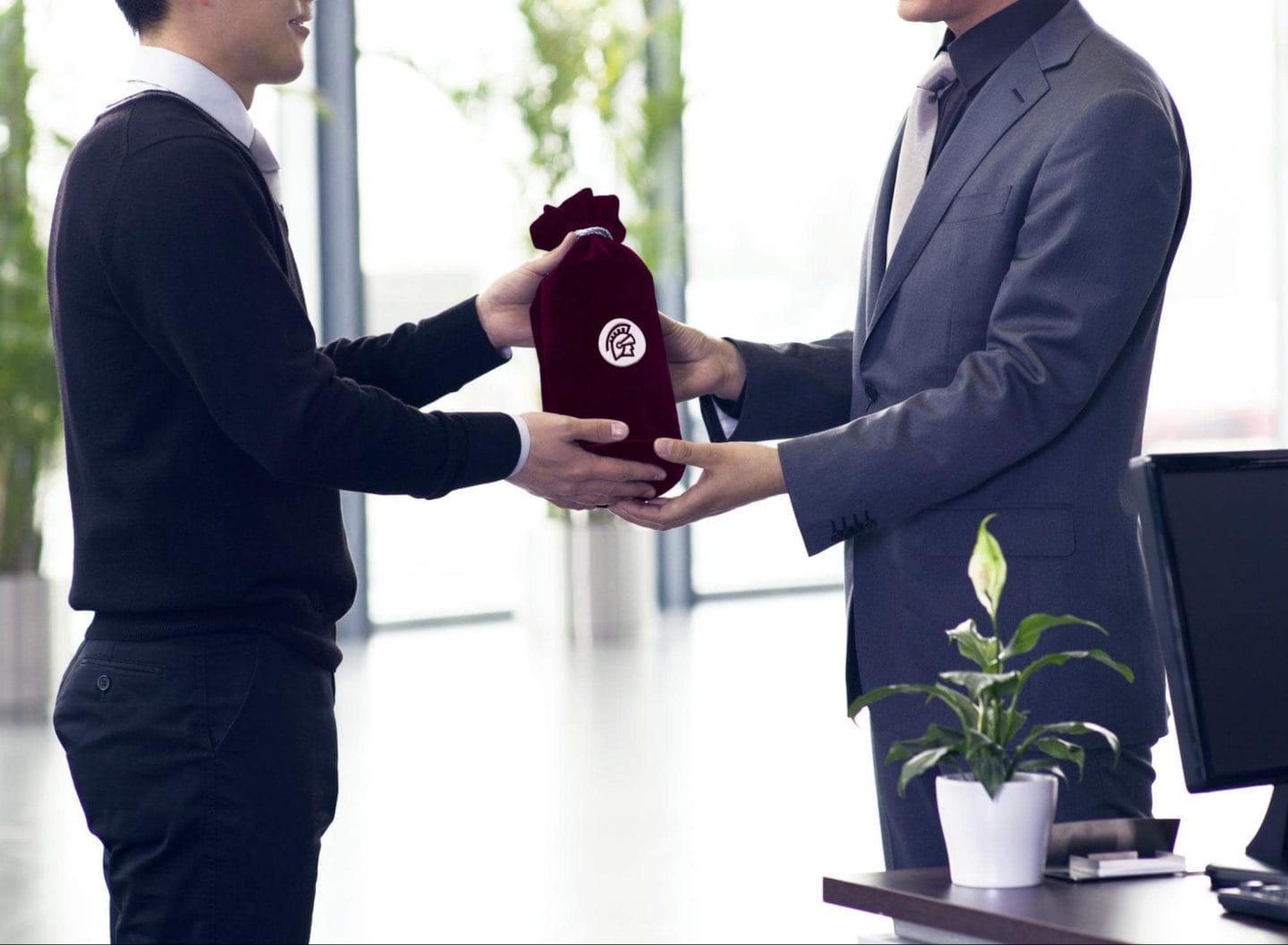 Two business men in an office. One is handing the other a gifted bottle of wine.