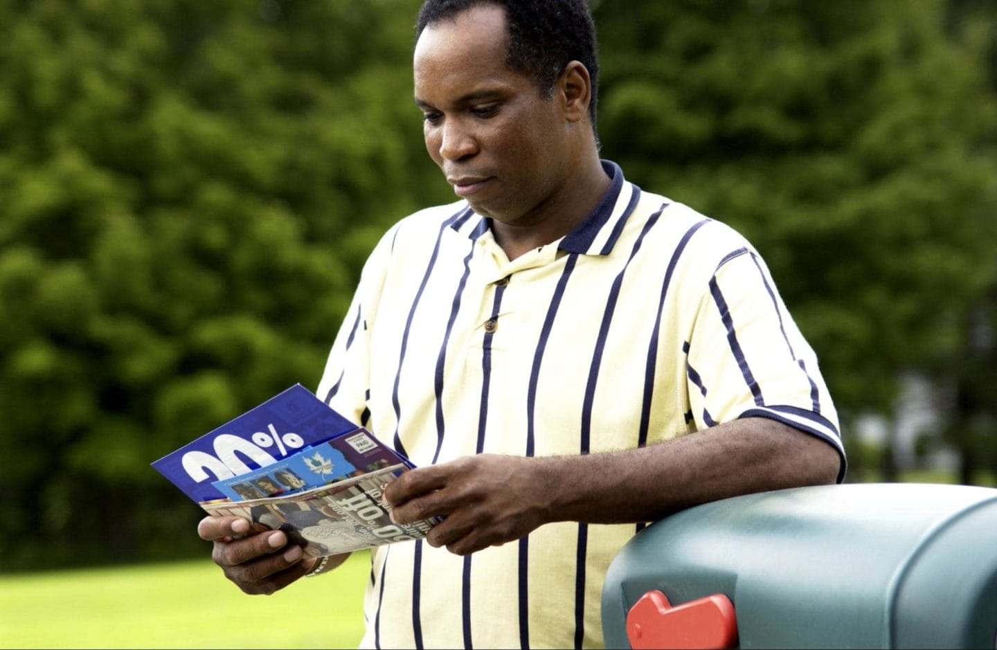 A man in a striped shirt getting mail from his mailbox and looking at the direct mail advertising pieces.