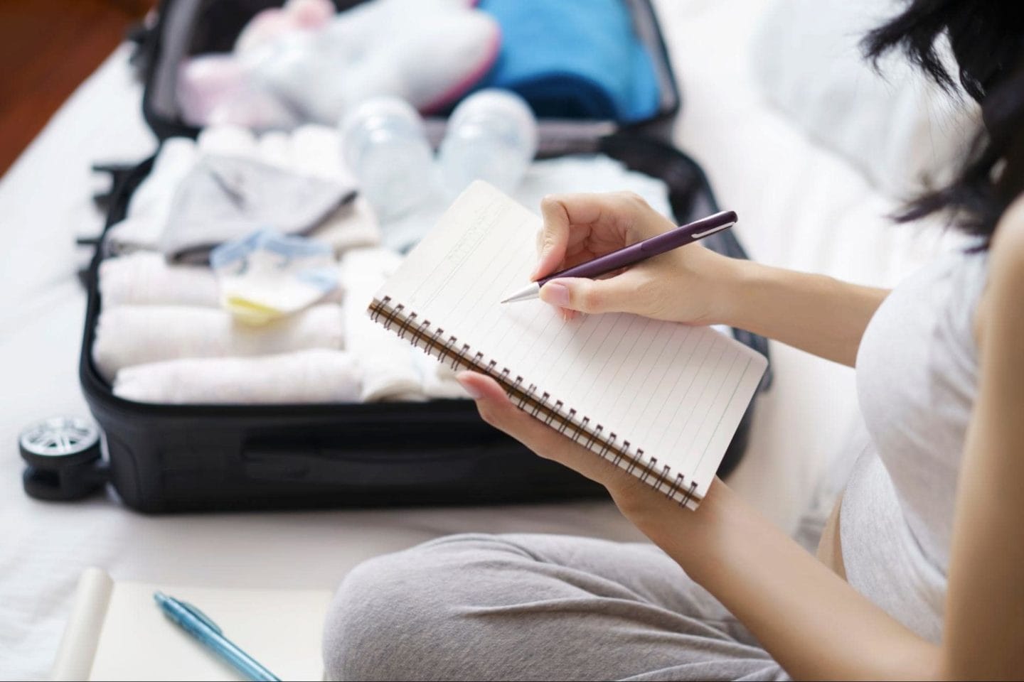 A woman sitting on her bed and writing in her notebook next to a packed suitcase.