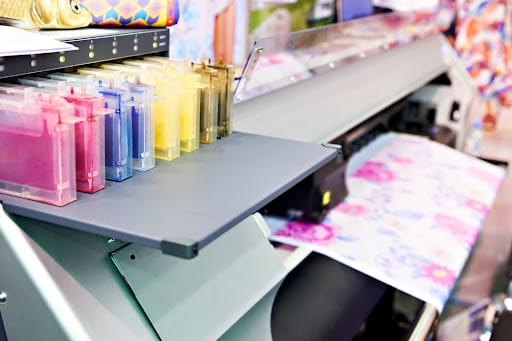 Printer ink is lined up sequentially by color from pink and blue to purple and yellow. There is a beautiful printed piece of paper with pinks and purples in the background.
