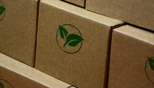 Cardboard boxes are stacked on top of each other in a pile. On the side of each box, a green leaf decal is printed in reference to going green.