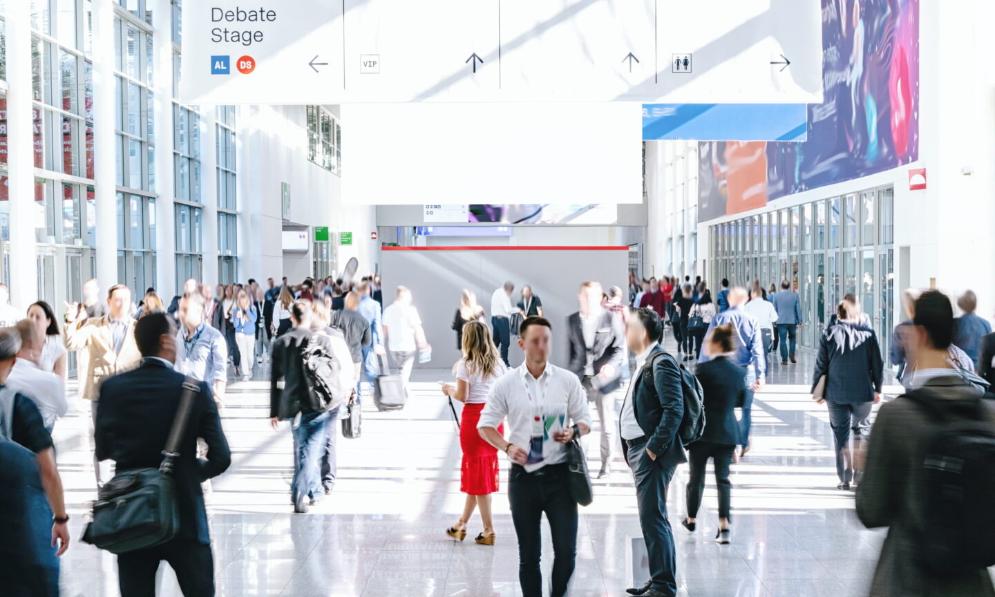 A bright, high-ceilinged convention center hallway filled with people walking between events. Overlaid at the top is a digital navigation banner showing a "Debate Stage" to the left, and icons with arrows pointing toward a VIP area, restrooms, and other directions. The scene captures the busy, professional atmosphere of a trade show or large conference.