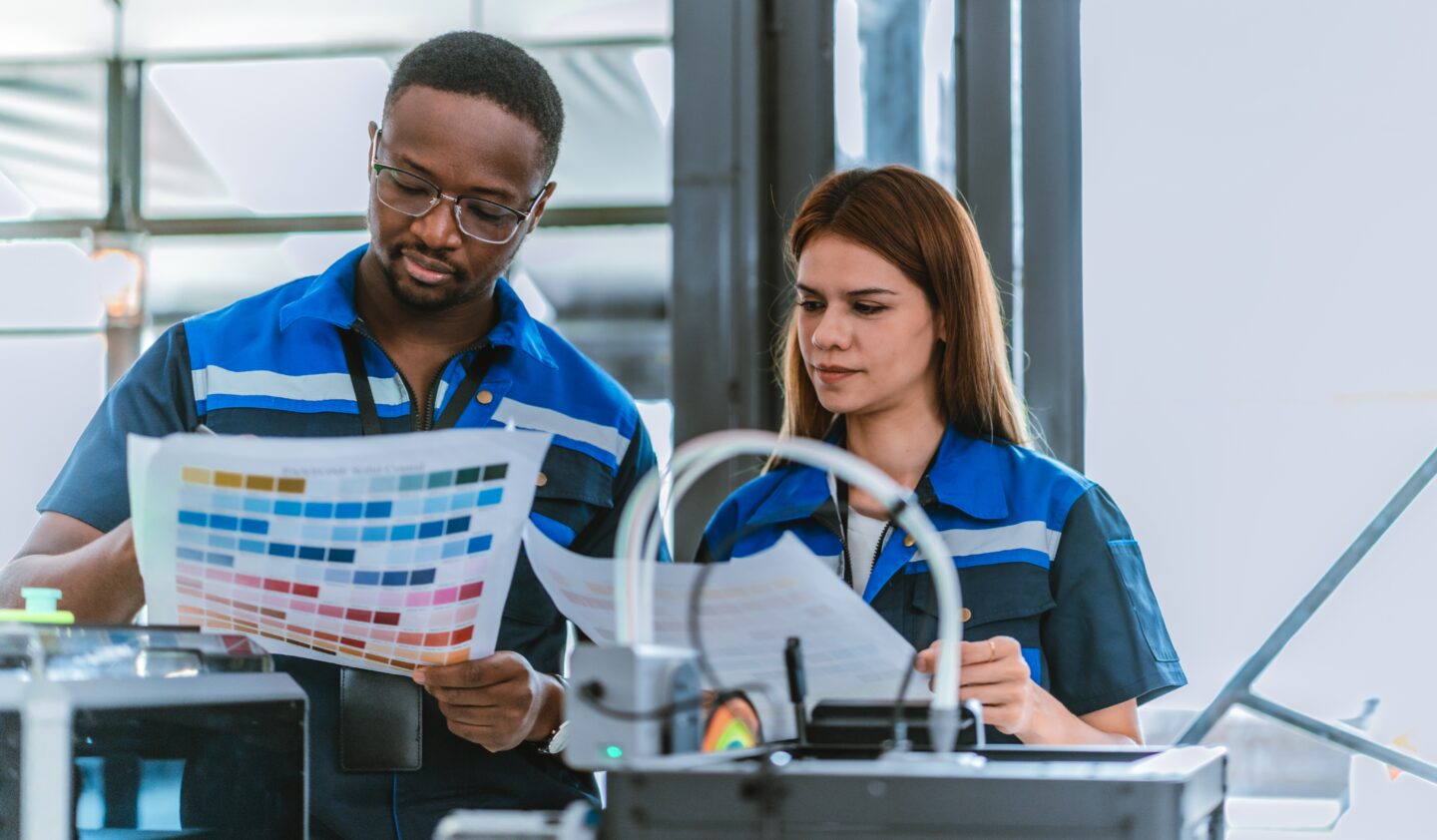 Two technicians in matching blue uniforms examine a printed sheet of color swatches while working at a machine, collaborating as a trusted label print partner to ensure accurate color matching.