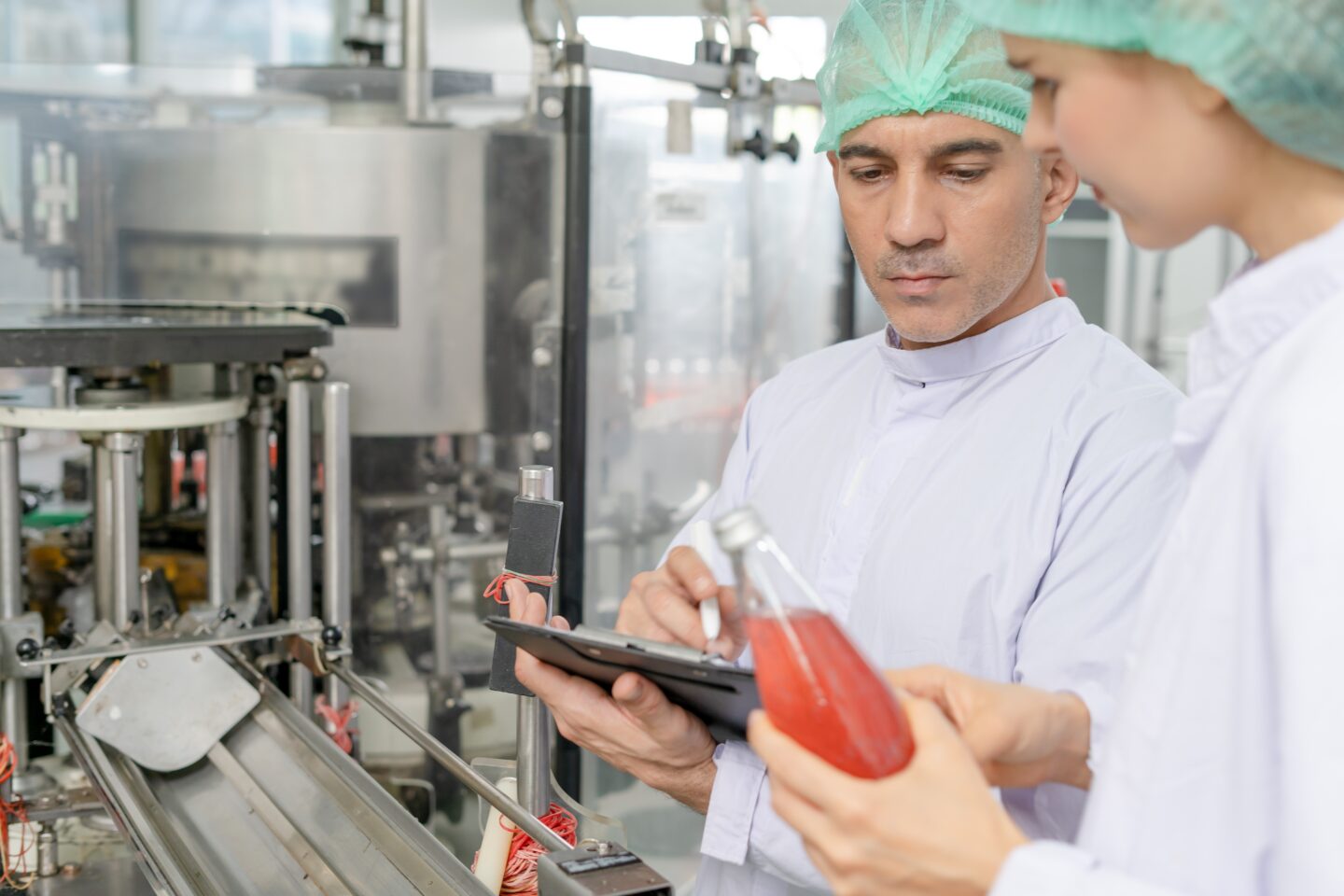 Two manufacturing workers in lab coats and hairnets perform a product packaging partner quality inspection by examining a bottled red beverage and taking notes on a clipboard next to an assembly line.