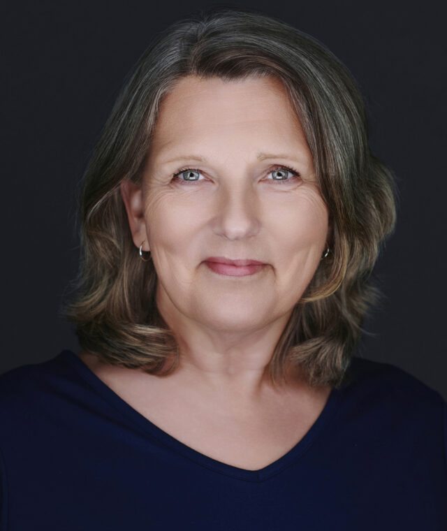 A studio headshot portrait of a mature woman with light skin, blue eyes, and shoulder-length brownish-gray hair. She is facing forward with a gentle, subtle smile against a solid black background, and is wearing a dark blue v-neck shirt.
