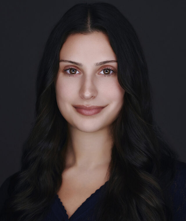 A studio headshot portrait of a young woman with a light complexion, brown eyes, and long, wavy dark hair parted down the middle. She is facing forward with a soft, gentle smile against a solid black background, and is wearing a dark blue top.